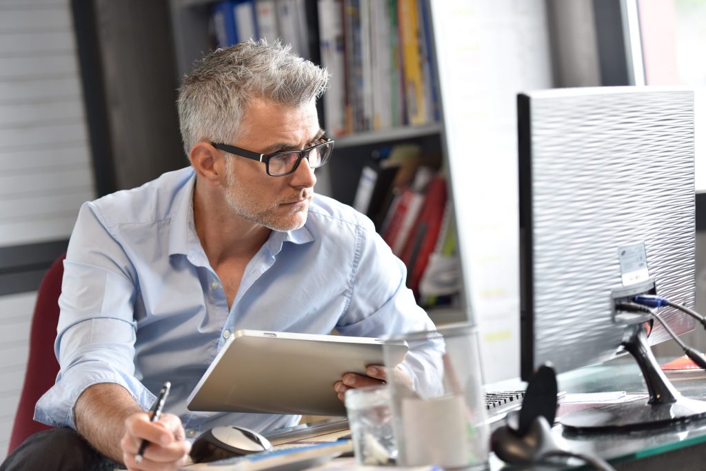 A man wearing glasses sits at a desk, writing with a pen on a piece of paper.